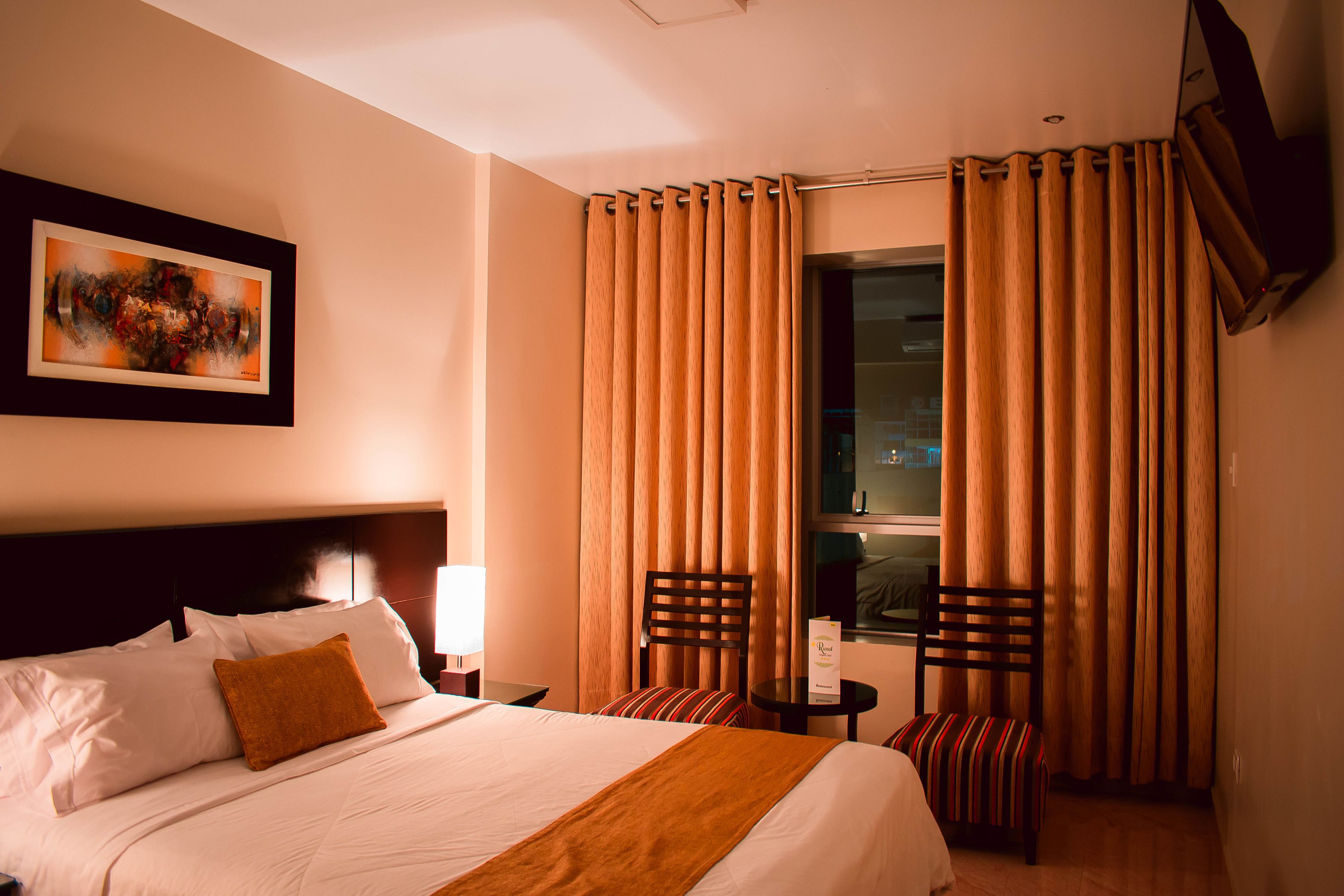Une chambre d’hôtel avec un grand lit, deux chaises, une petite table et une fenêtre avec des rideaux.