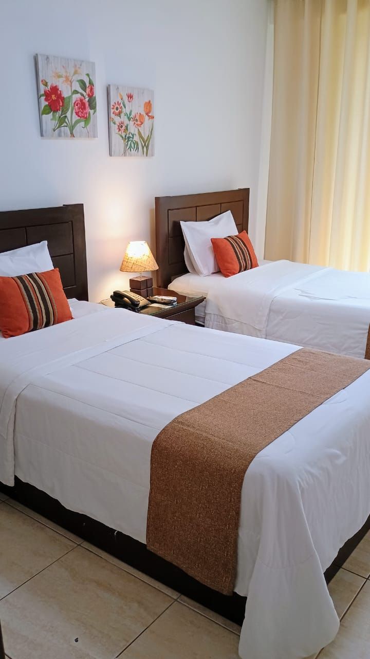 Habitación de hotel con dos camas, una mesita de noche, una lámpara y dos cuadros con motivos florales en la pared.