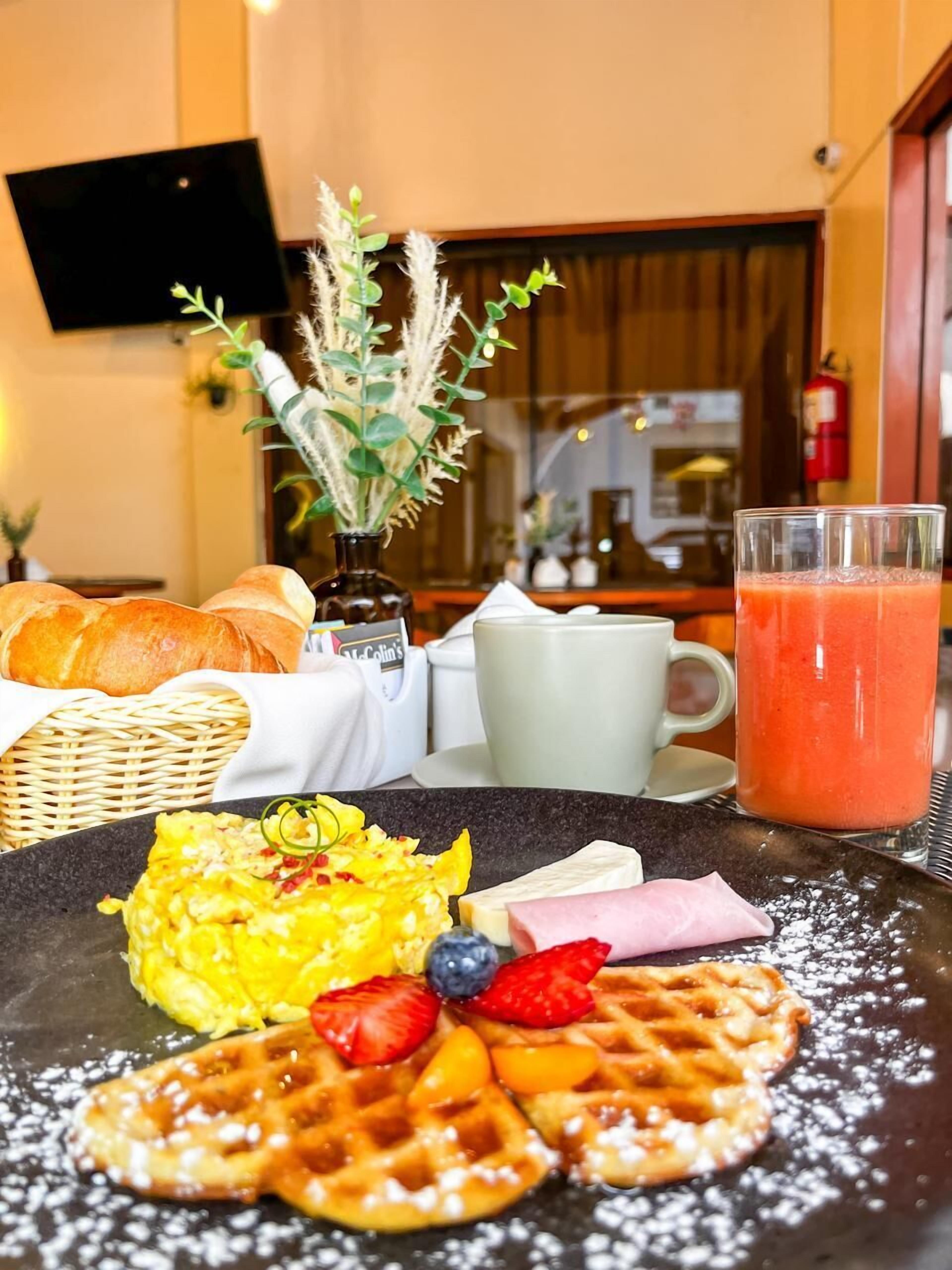 En frukostuppläggning med våfflor, äggröra, ost, skinka, jordgubbar och blåbär på en tallrik, ett glas apelsinjuice och en vas med torkade växter.
