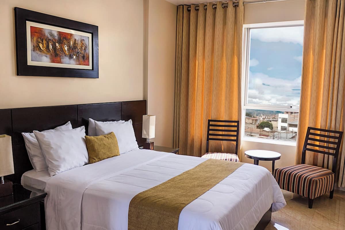 A hotel room with a large bed, a headboard, a nightstand, a lamp, a chair, a small table, a window with curtains, and a painting on the wall.