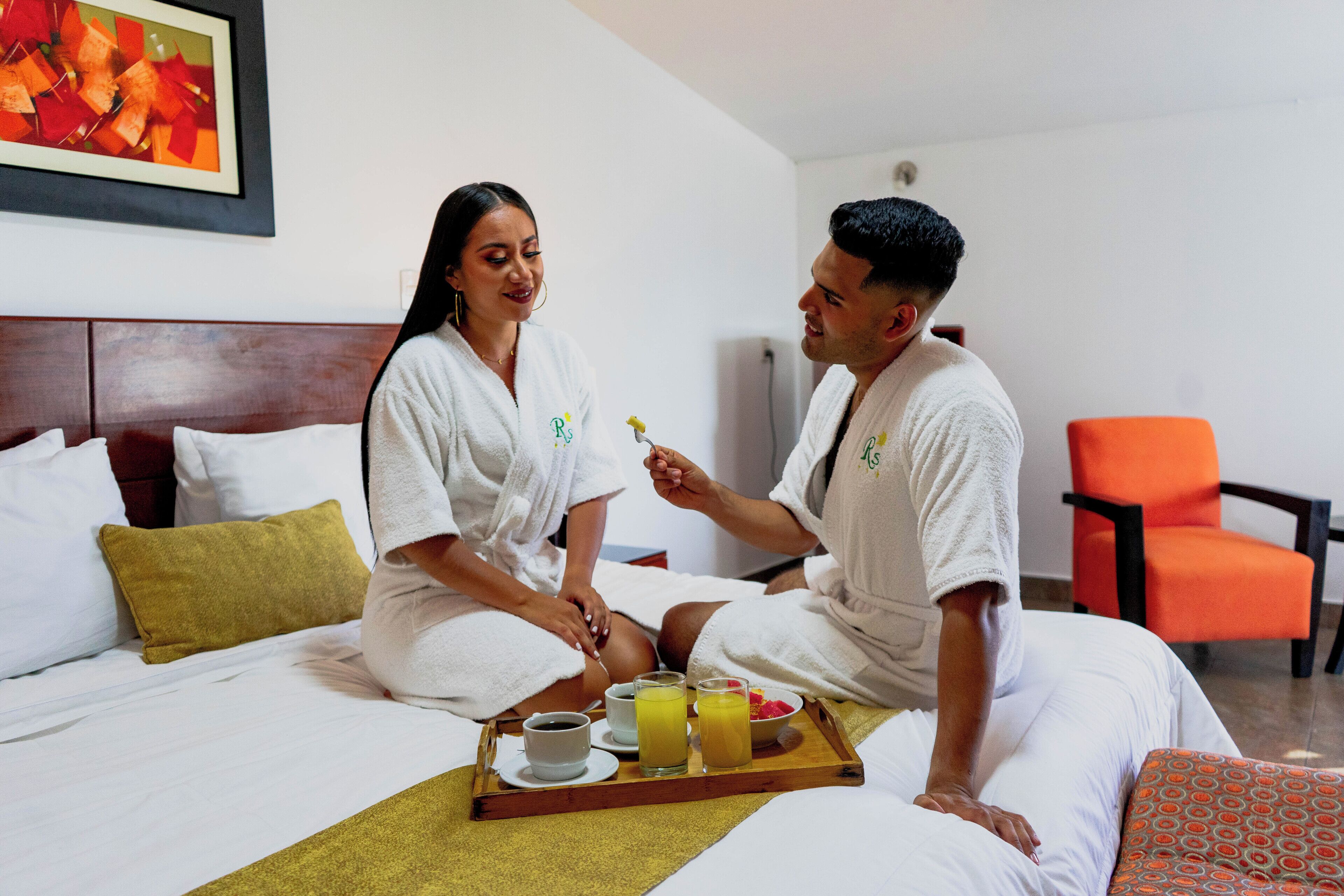 A couple in white robes sitting on a bed with a tray of breakfast items including coffee, juice, and a bowl of fruit.