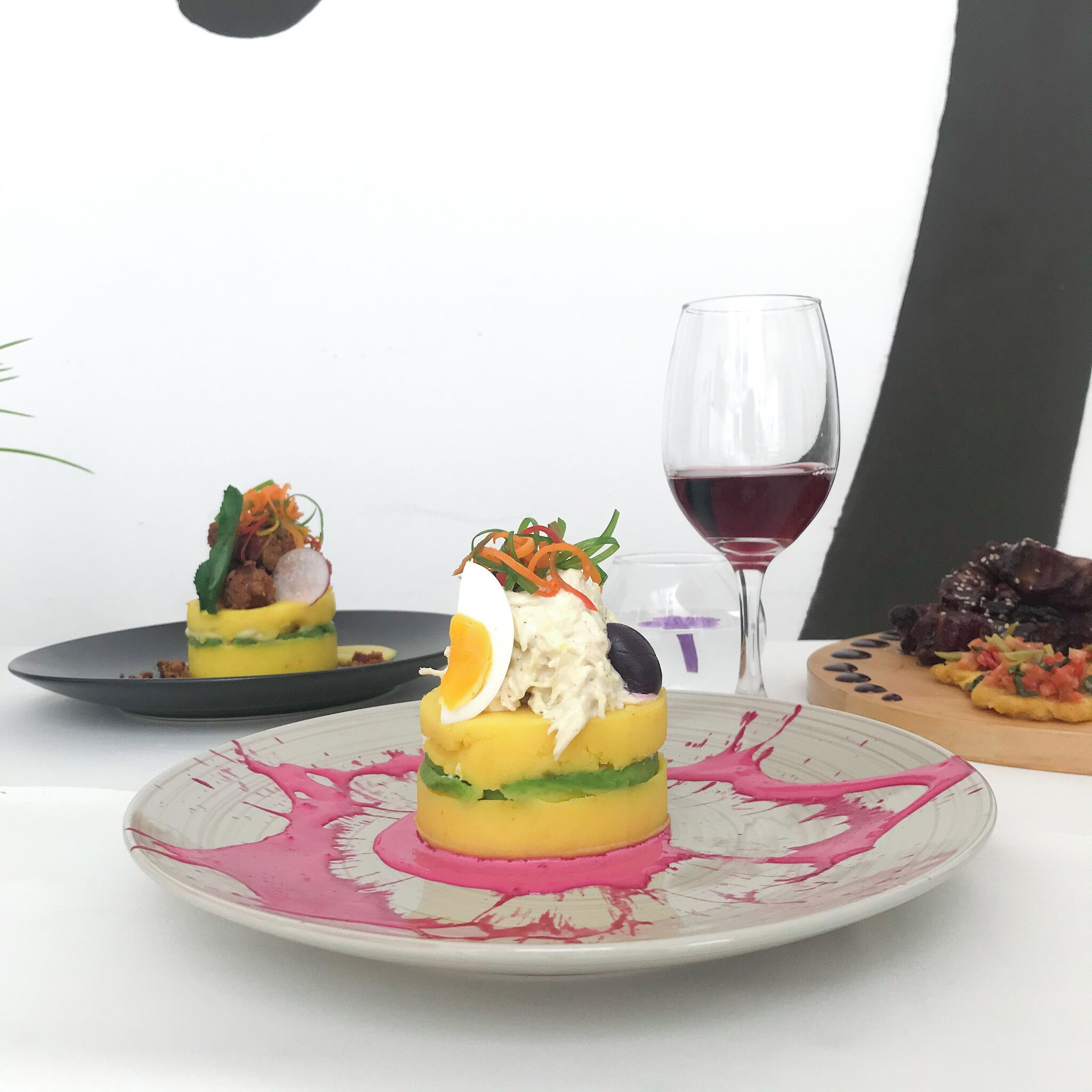 A dessert on a plate with a pink and white pattern, a glass of red wine, and a wooden board with another dish.
