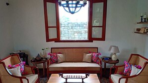 Een kamer met een bank, twee fauteuils, een salontafel, een kroonluchter, een raam met geopende luiken en een stenen muur buiten.