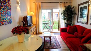 Un salon avec un canapé rouge, une petite table sur laquelle est posé un vase de roses rouges, un téléviseur sur un meuble en bois, et un balcon avec une table et des chaises.