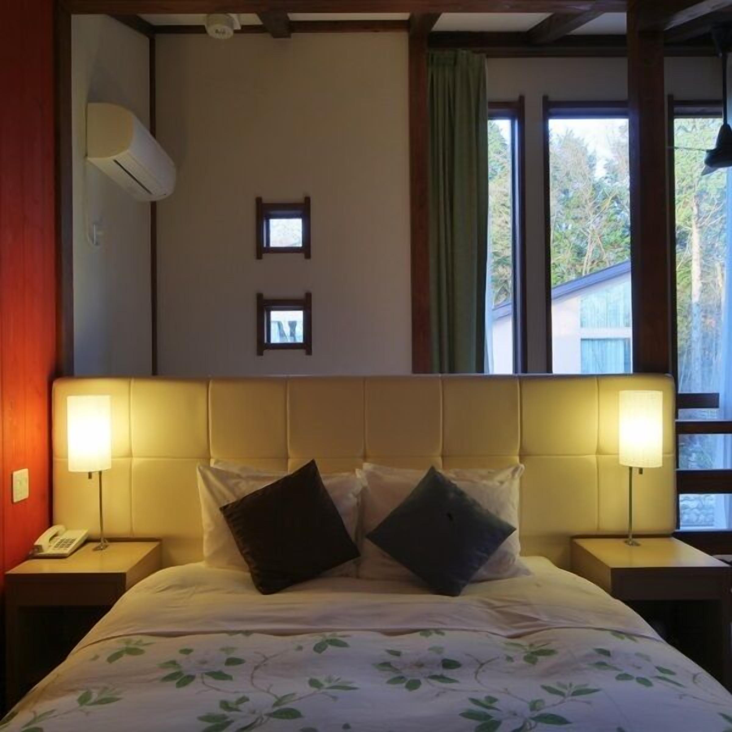 Ein Schlafzimmer mit Kopfteil, zwei Nachttischlampen, einem Telefon, einem Bett mit Kissen, zwei kleinen Bildern an der Wand und einem Fenster mit Vorhängen.