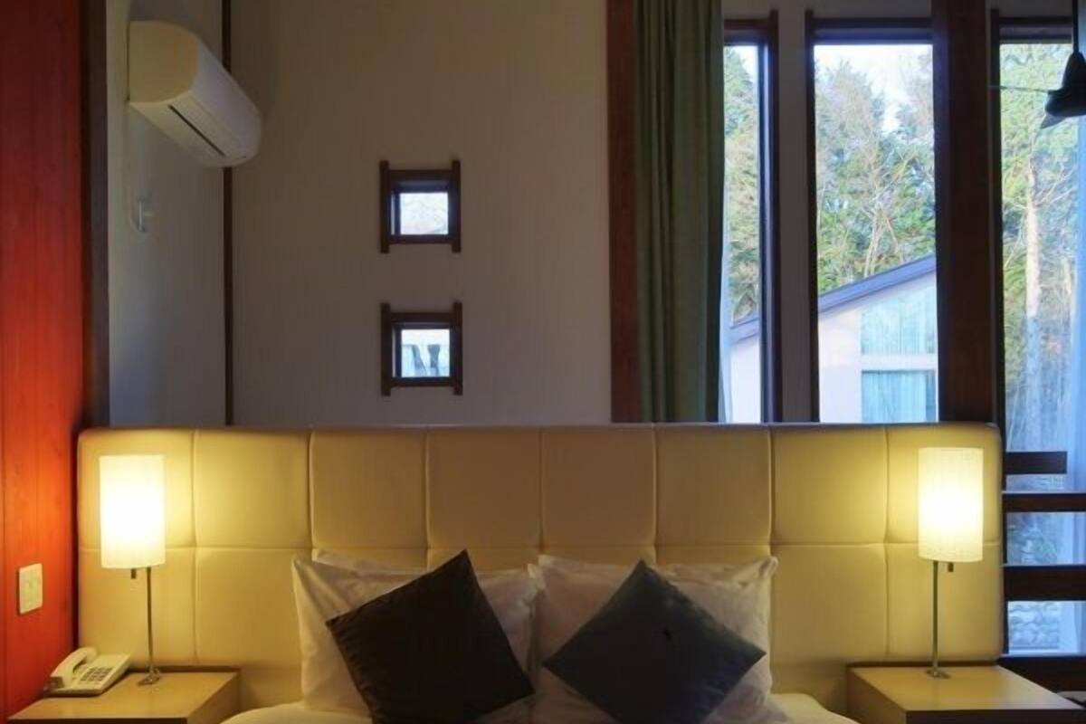 A bedroom with a headboard, two bedside lamps, a telephone, a bed with pillows, two small wall-mounted pictures, and a window with curtains.