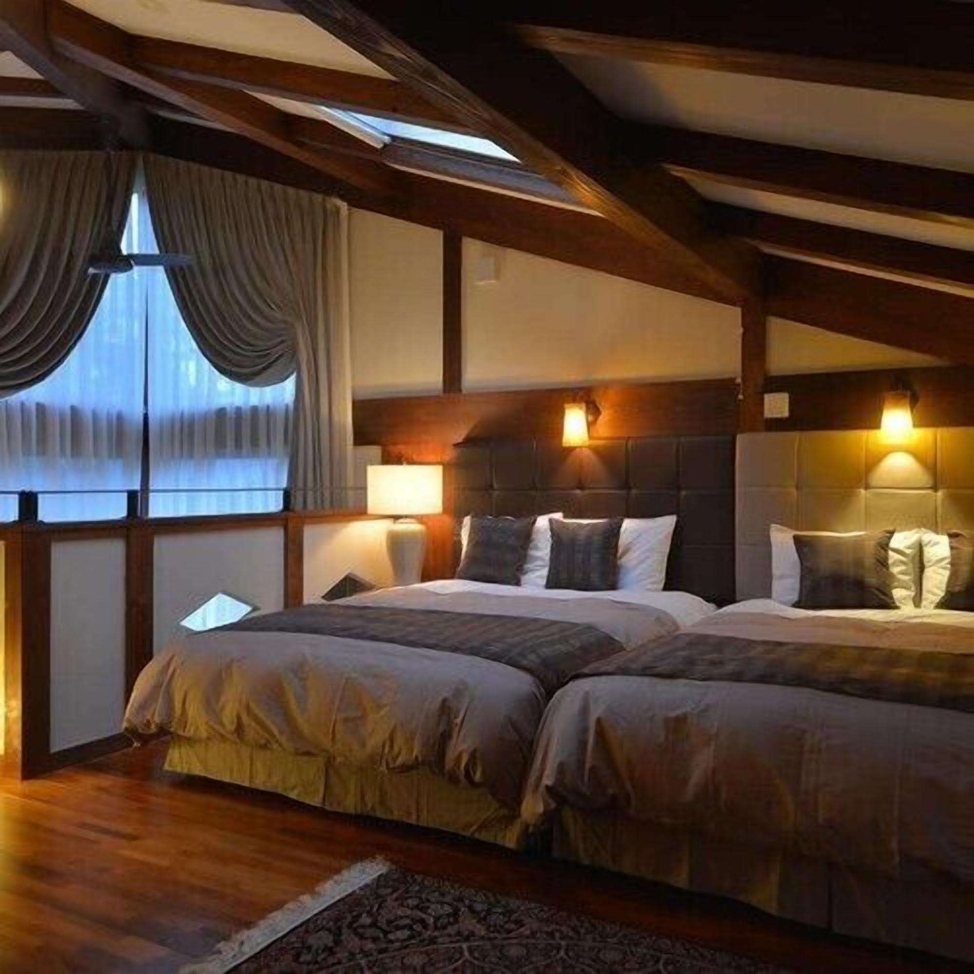 Ein Zimmer mit zwei Betten, Holzbalken, einem großen Fenster mit Vorhängen und Wandlampen.