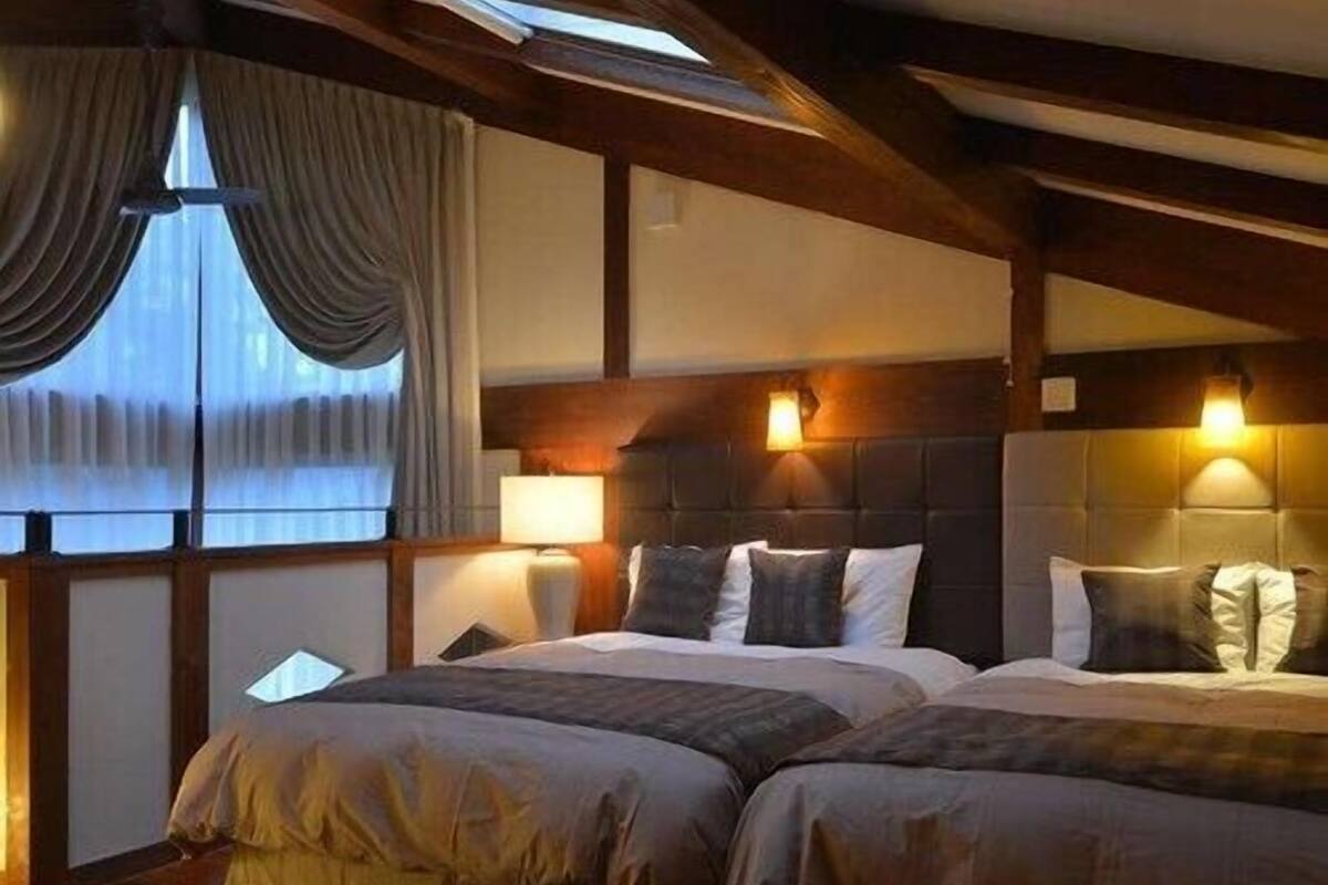 A room with two beds, wooden beams, a large window with curtains, and wall-mounted lamps.
