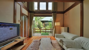 A room with a wooden coffee table, a sofa, a TV, and a view of a green outdoor area through a glass door. - One More Time Heart (Nikko)