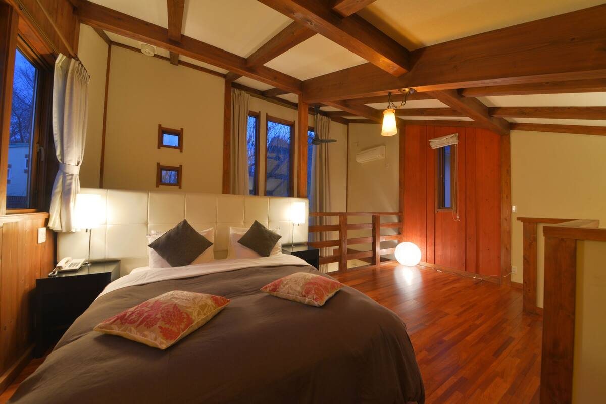A bedroom with a wooden ceiling, a bed with multiple pillows, a nightstand with a phone, and a window with curtains.