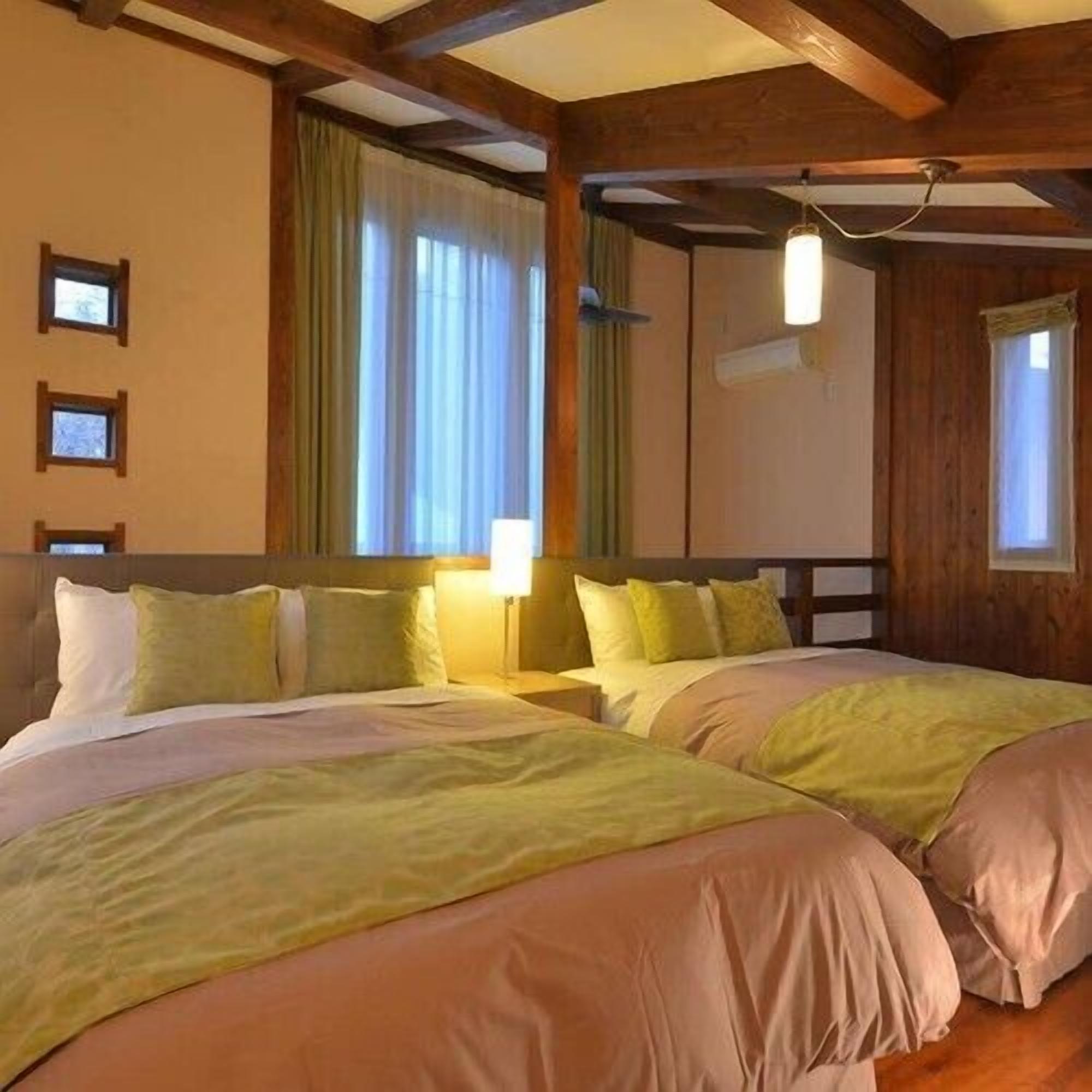 Ein Zimmer mit zwei Betten, Holzbalken, einem fenster mit Vorhängen und einer Wandleuchte.