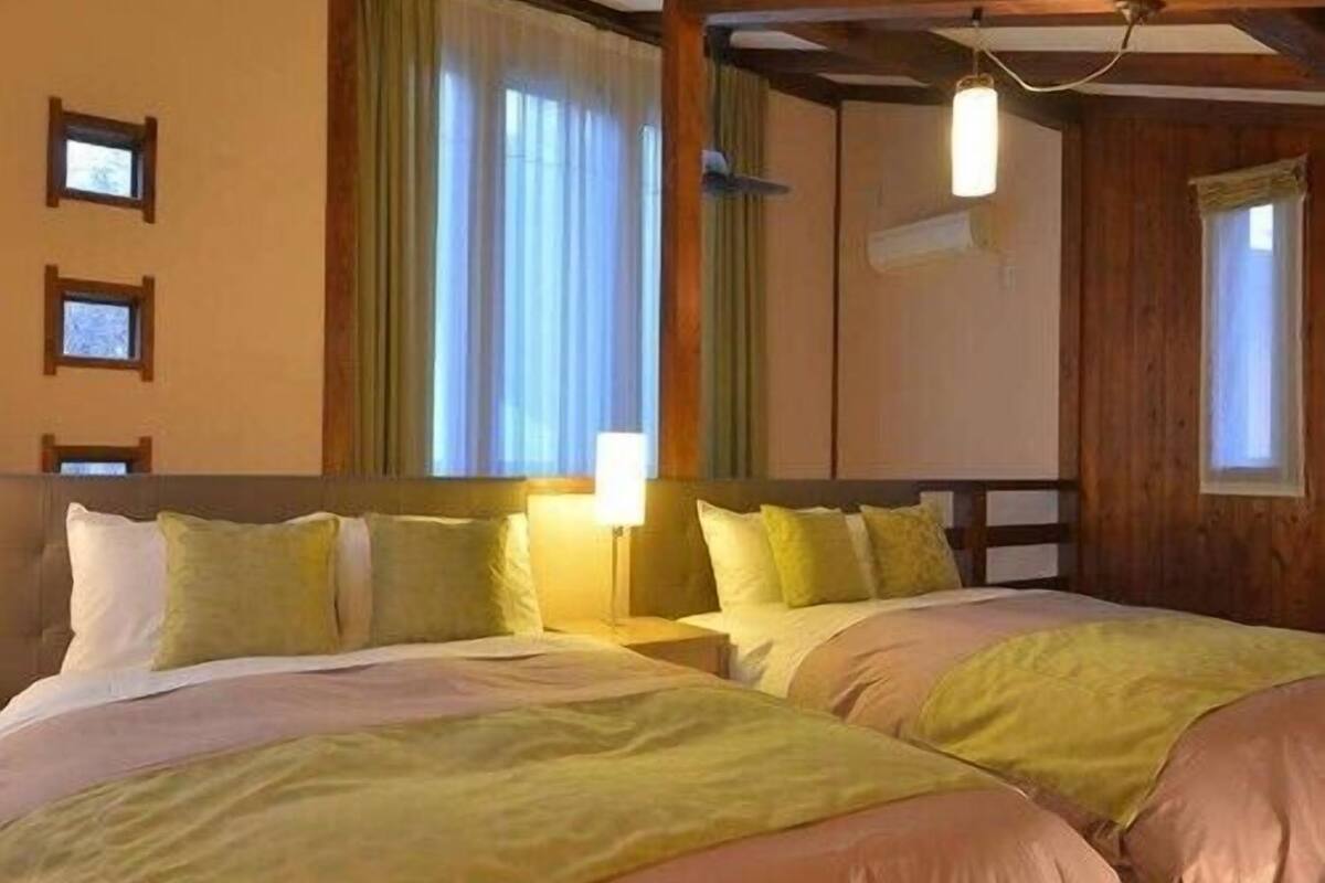 A room with two beds, wooden beams, a window with curtains, and a wall-mounted light fixture.