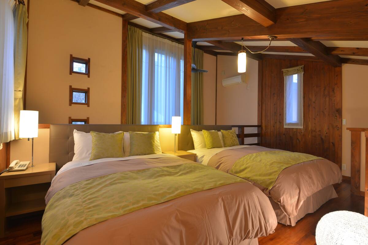 A double room with two beds, wooden beams, a telephone on the nightstand, and a window with curtains.