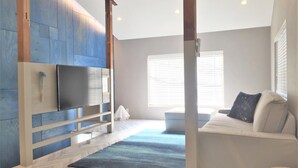 Une pièce moderne avec un parquet en chevrons, un mur d’accent bleu, un téléviseur à écran plat fixé sur un panneau en bois, un canapé avec un coussin et une fenêtre avec des stores.