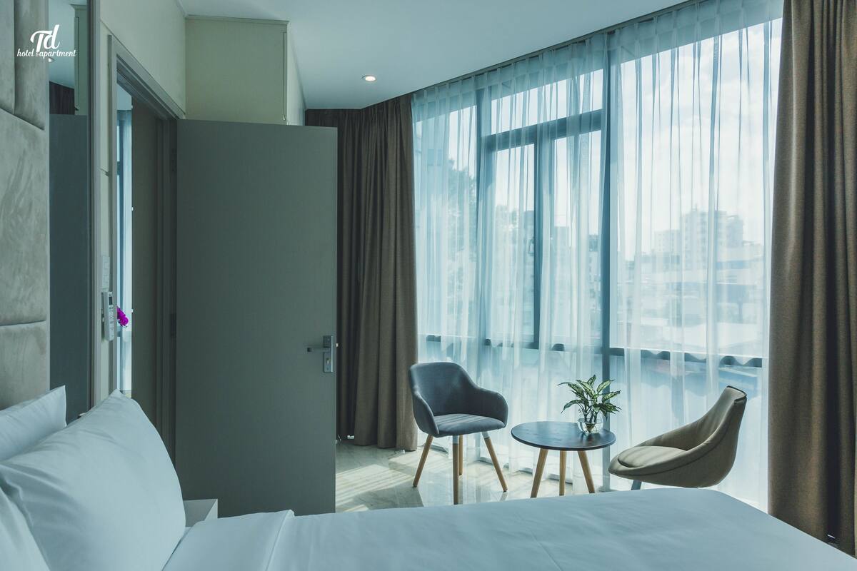 A hotel room with a bed, two chairs, a small table, and a large window with curtains.