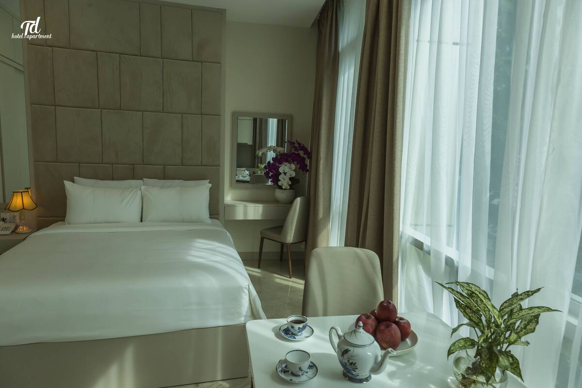A hotel room with a large bed, a bedside table with a teapot and cups, a chair, a desk with a mirror, and a window with curtains.