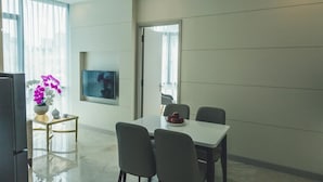 Habitación de hotel moderna con zona de comedor que incluye una mesa blanca y cuatro sillas, un televisor de pantalla plana en la pared y un ventanal con cortinas traslúcidas.