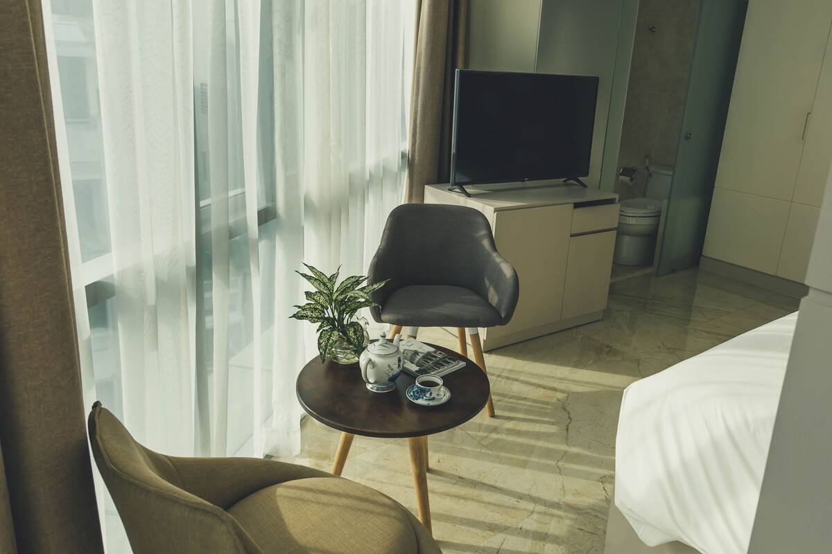 A hotel room with a bed, a TV, a small table with a plant and a cup, and a window with curtains.