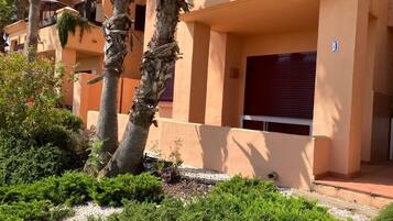 Un bâtiment à la façade beige, avec des palmiers et une fenêtre à volets rouges.