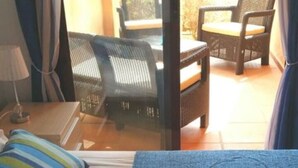 A hotel room with a bed, a blue blanket, a striped pillow, a nightstand with a lamp, and a view of an outdoor seating area with chairs and a table.