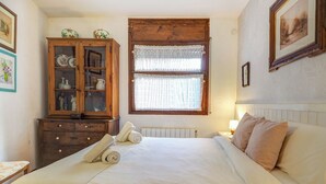 Une chambre à coucher avec un grand lit, une armoire en bois aux portes vitrées, une fenêtre ornée de rideaux en dentelle et un radiateur.