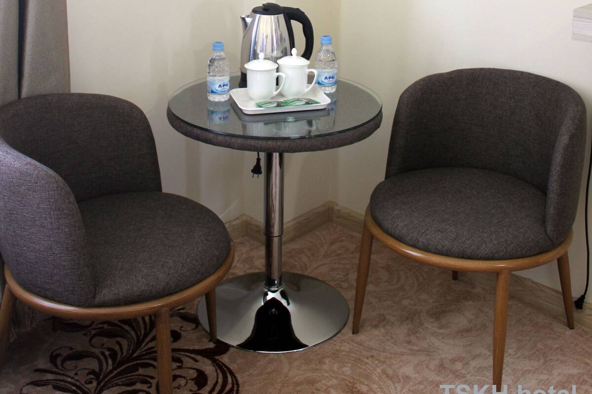 A corner room with two grey upholstered chairs, a small round table with a kettle, cups, and a water bottle.