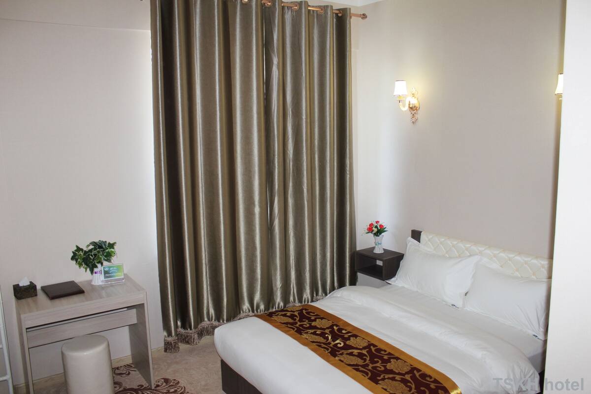 A hotel room with a bed, a desk, a chair, a nightstand, a lamp, and a curtain.