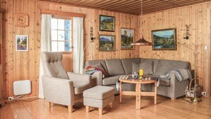 A cozy living room with wooden walls, a large window, a grey sofa, a chair, and a small table with a candle.
