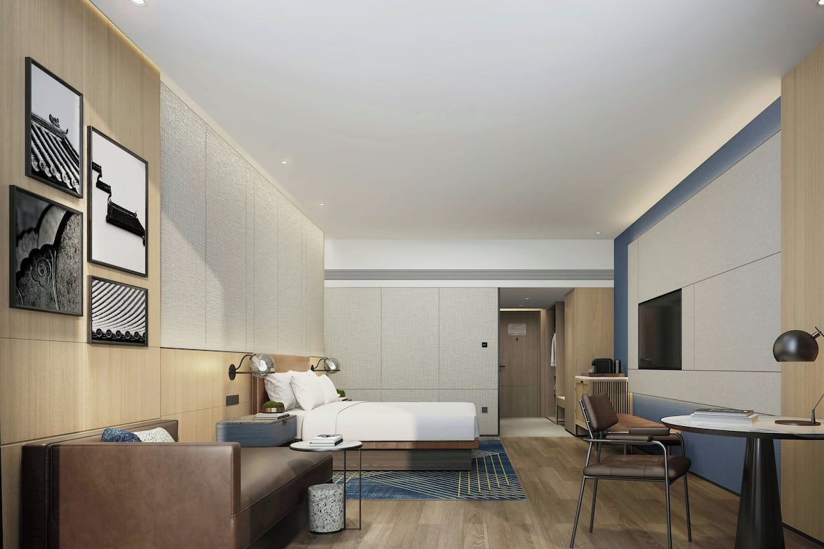 A modern hotel room with a bed, a sofa, a desk, and a chair. There are framed pictures on the wall and a window in the room.