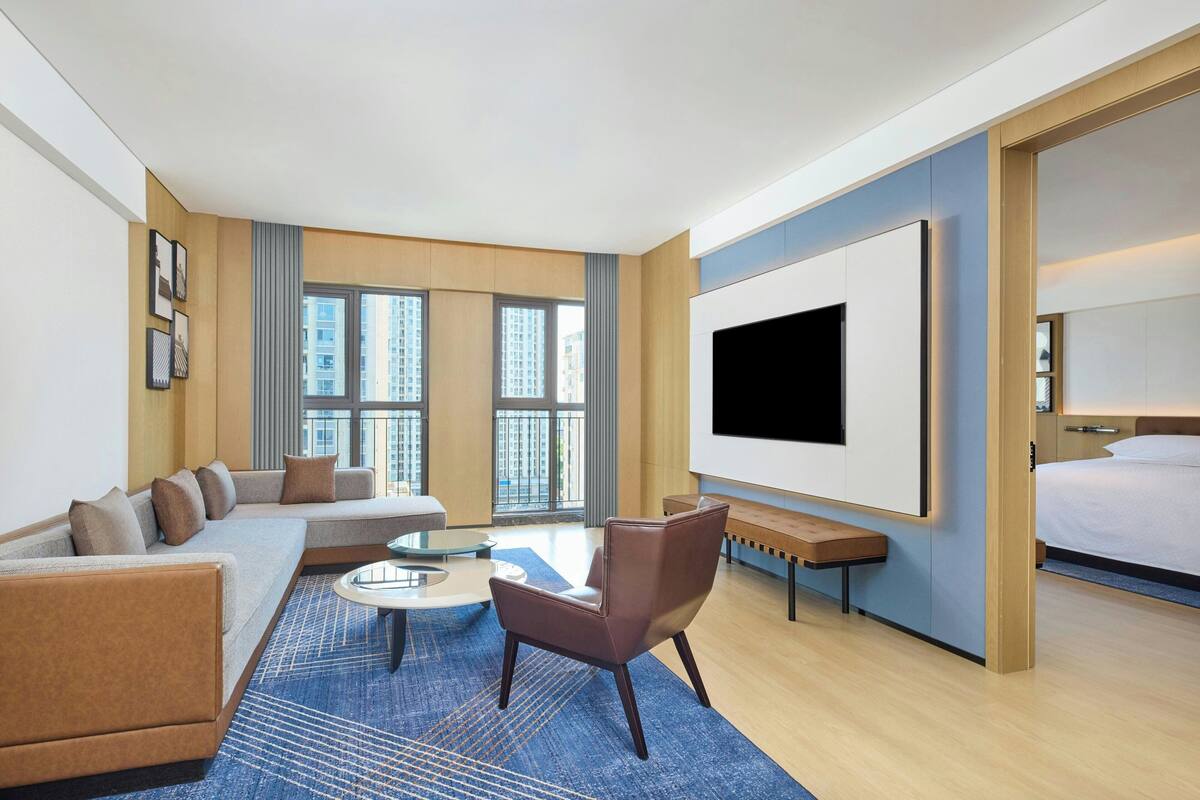 A modern hotel room with a large bed, a sofa, a chair, a coffee table, and a TV.