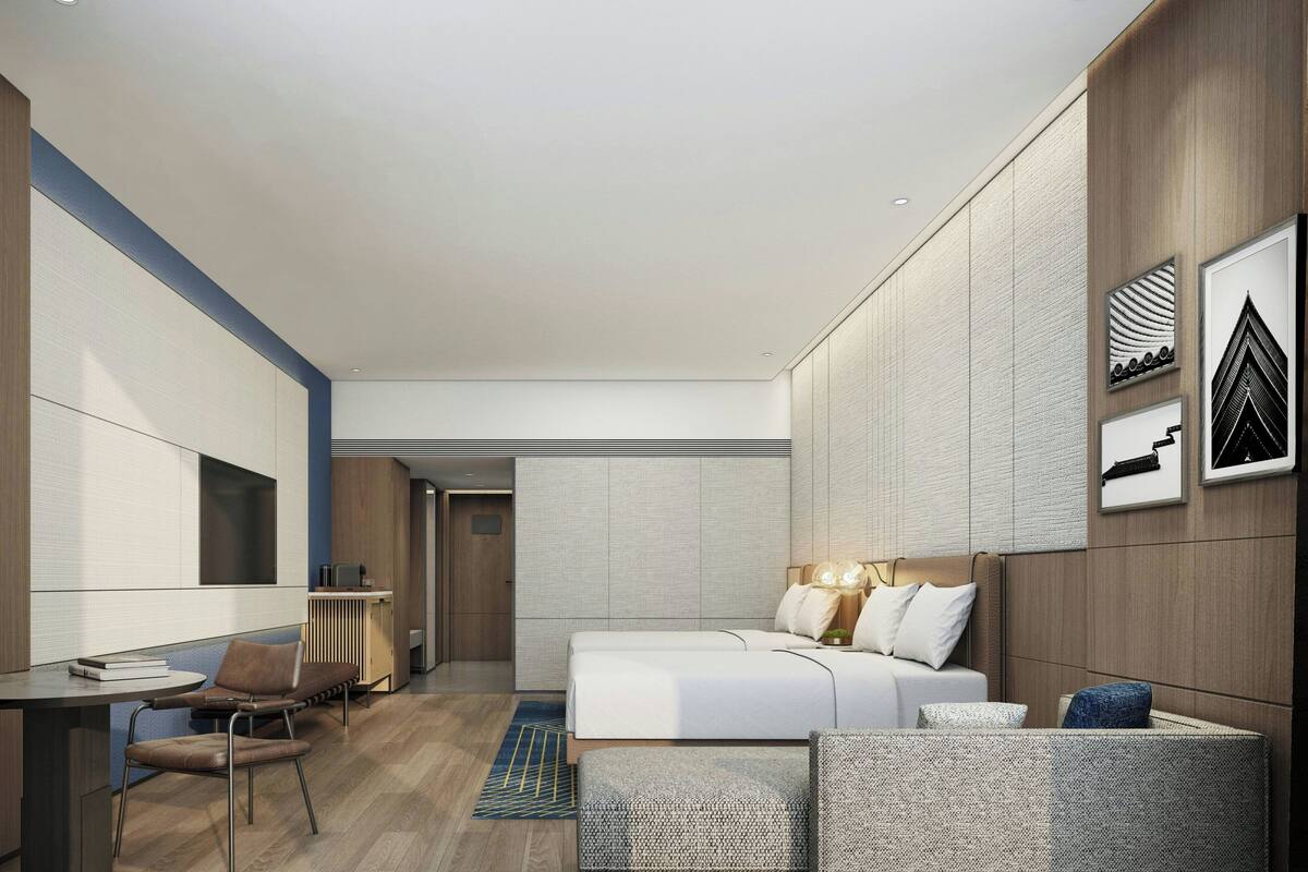 A modern hotel room with a bed, a desk, and a chair. There are framed pictures on the wall and a small table with a lamp.