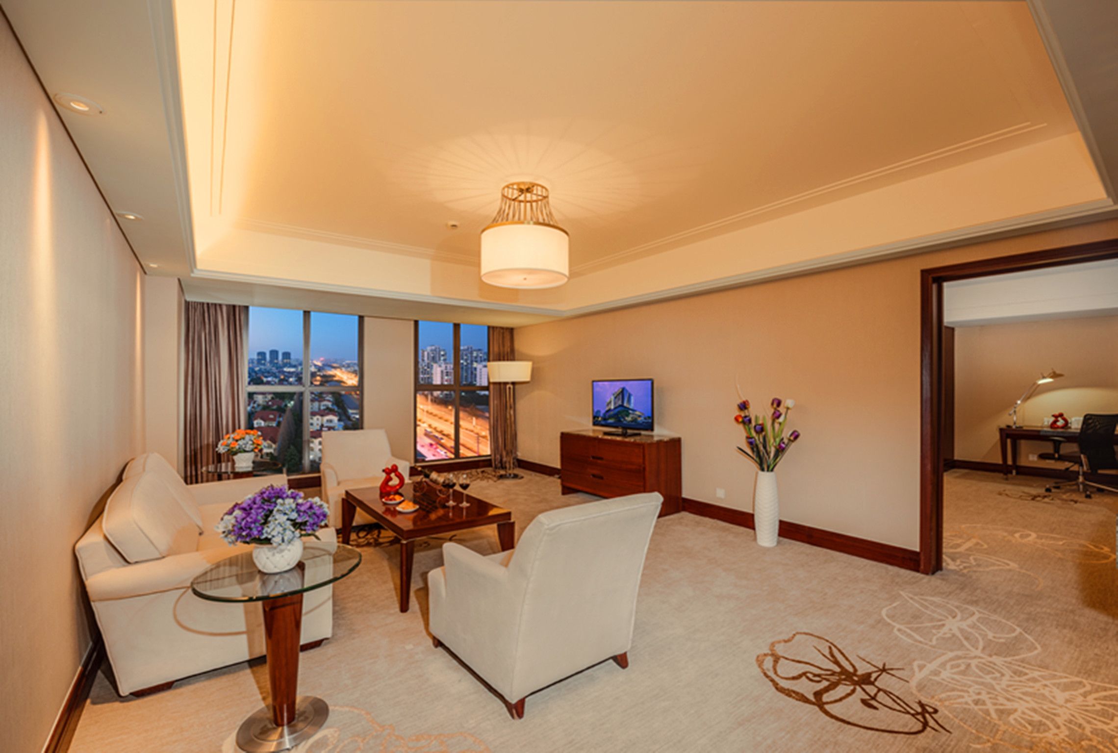 A modern hotel room with a sofa, armchair, glass coffee table, and a television.