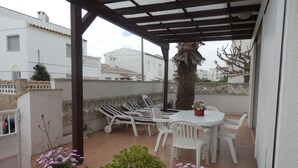 Eine überdachte Terrasse mit weißen Stühlen und einem Tisch, umgeben von Kübelpflanzen und Blumen.