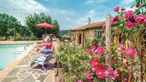Una piscina de piedra con sillones de descanso y una sombrilla roja, rodeada de exuberante vegetación y rosales florecientes.