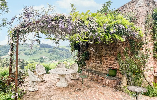 Un espace extérieur aménagé dans un style rustique, avec des murs en pierre, une vue sur des collines verdoyantes, ainsi qu’une table et des chaises en pierre.