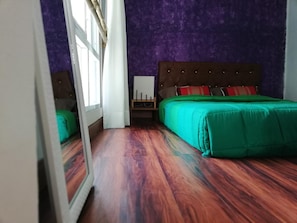 A bedroom with a wooden floor, a bed with green bedding, purple walls, and a window with curtains.