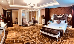 Une chambre spacieuse avec un grand lit, un coin salon comprenant un fauteuil et une table, une coiffeuse avec un miroir et un lustre.