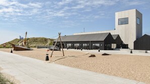 Un edificio moderno con techo plano y ventanas amplias, ubicado en una zona arenosa con un camino pavimentado.