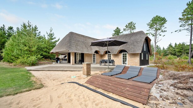 Una casa de playa con techo de paja, mobiliario exterior y un patio arenoso.