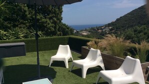 Eine Terrasse mit weißen Plastikstühlen und einem Tisch, umgeben von grünem Kunstgrün mit Blick auf eine Hügelkette und den Ozean.