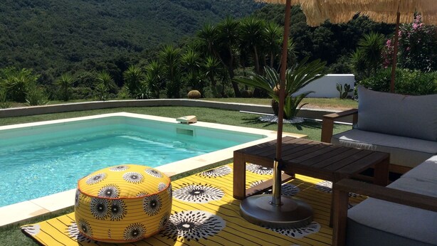 Ein Poolbereich mit Holzterrasse, einem gelb-schwarz gemusterten Pouf, einem Holztisch und einem Reetdachschirm.