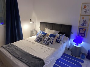 A neatly made bed with white bedding and blue coral-patterned pillows, a bedside lamp, and a wall-mounted picture frame.