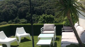 Un espace extérieur aménagé avec des fauteuils de jardin blancs et une table, entouré de haies bien entretenues et offrant une vue sur une colline boisée.