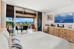 A hotel room with a bed, a TV, a dining area with chairs, and a view of the ocean.