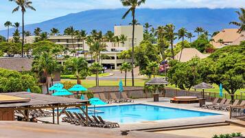 A resort pool area with lounge chairs and umbrellas, surrounded by palm trees and a view of a mountain range.