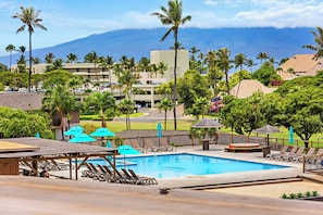 Un área de piscina en un resort con sillones de descanso y sombrillas, rodeada de palmeras y vistas a una cadena montañosa.