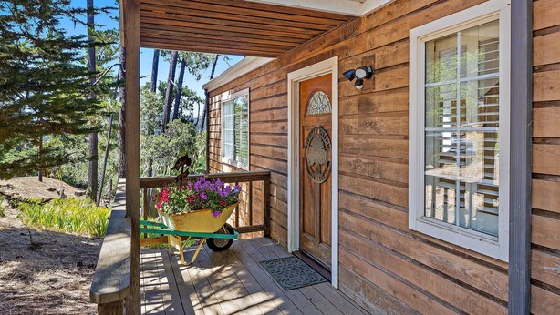 Une cabane en bois avec un porche, une brouette remplie de fleurs et une porte ornée d’un emblème circulaire.
