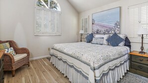 Une chambre à coucher avec un grand lit, une chaise en osier, une commode en bois et une fenêtre avec des volets.