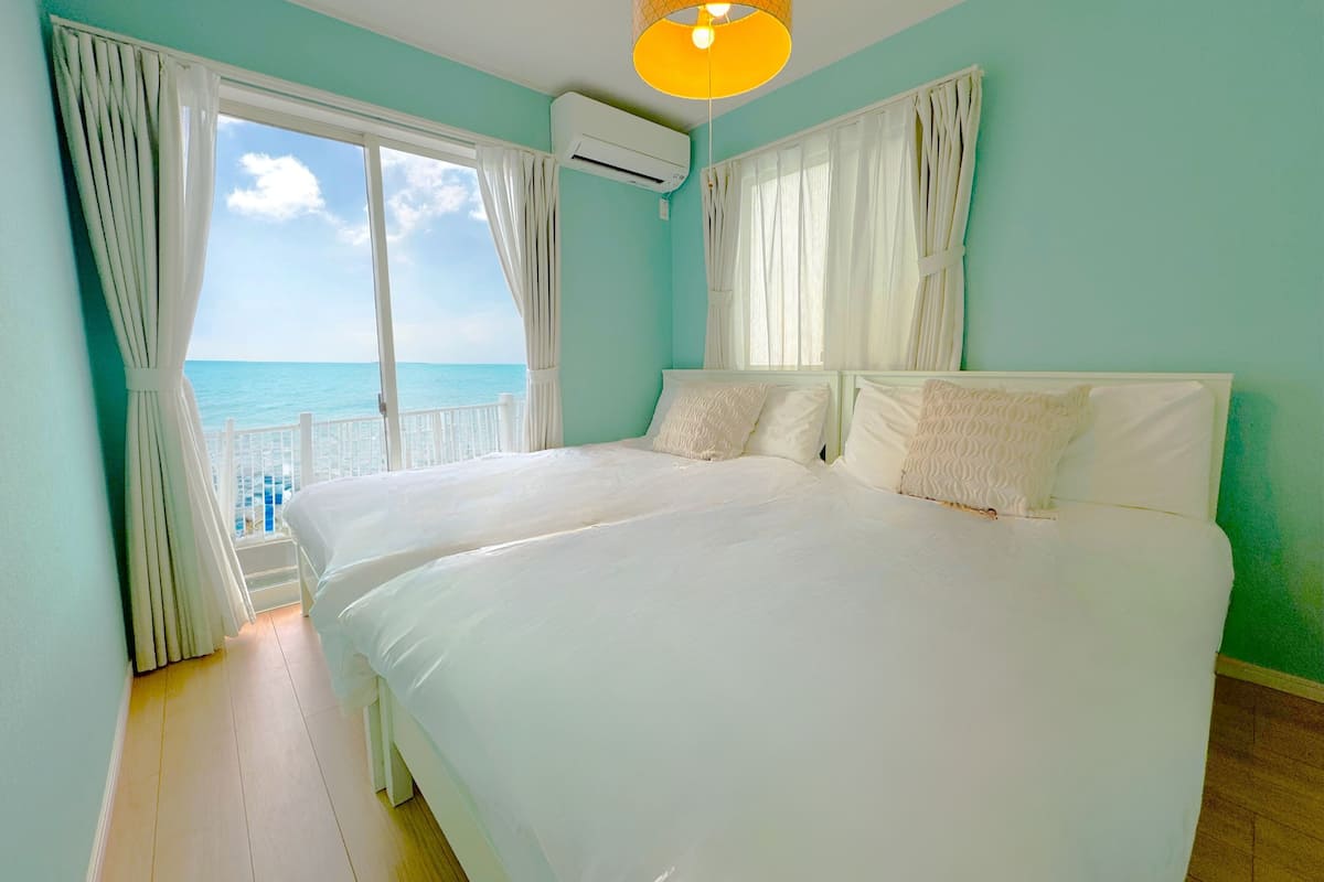 A bedroom with a large bed, white curtains, an air conditioning unit, and a view of the sea.