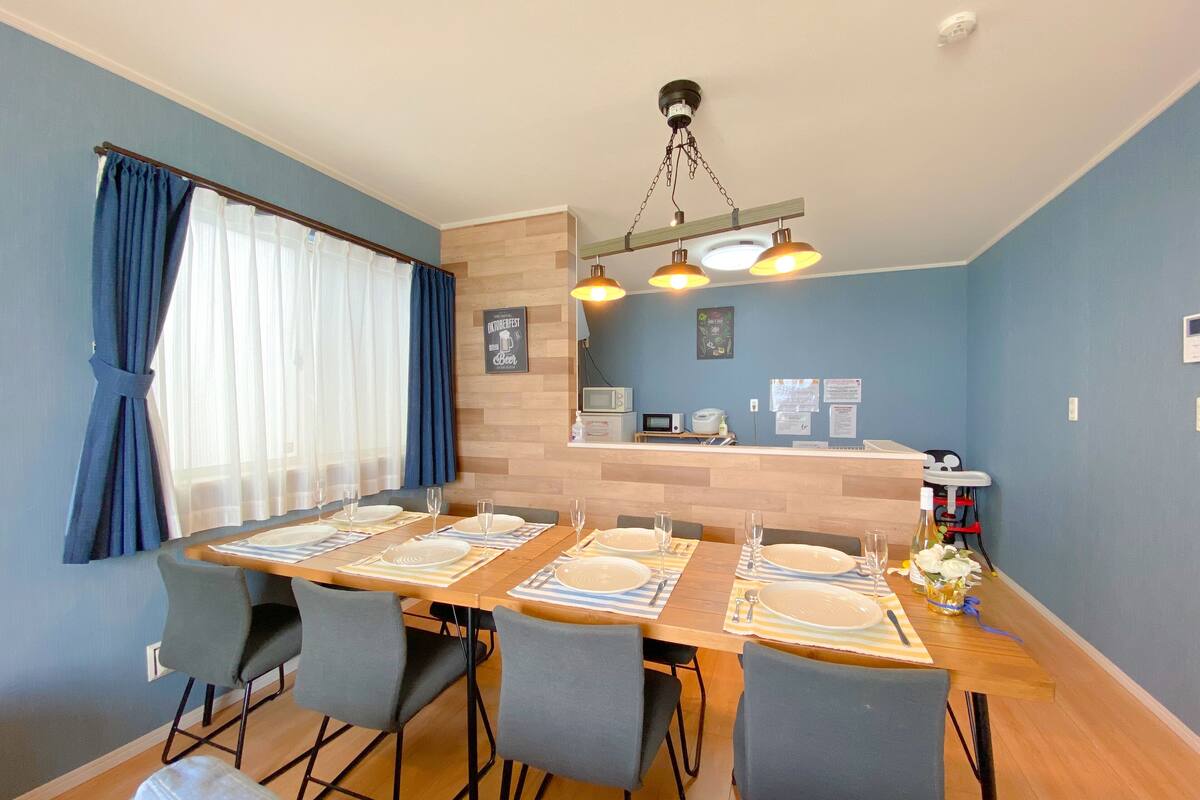 A dining area with a wooden table set for six, six chairs, and a kitchen area in the background.